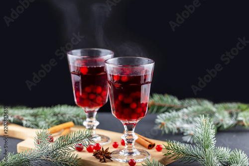 Two stemmed glasses hold deep red spiced drinks beside evergreen branches, berries, cinnamon sticks, and star anise on wooden tabletop, with subtle steam rising against dark background