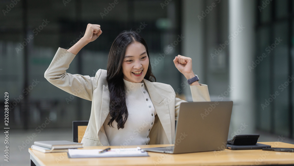 Fototapeta premium Excited woman cheering with fists raised, achieving business success while working on a laptop in a modern office