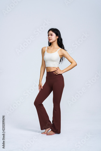 Young East Asian woman in white sports top and brown high-waisted yoga flare pants posing in plain light grey studio 
