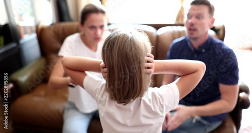 In a warm living room, a young girl holds her ears, overwhelmed by her parents' animated conversation. They appear upset, discussing matters that trouble the family dynamic.