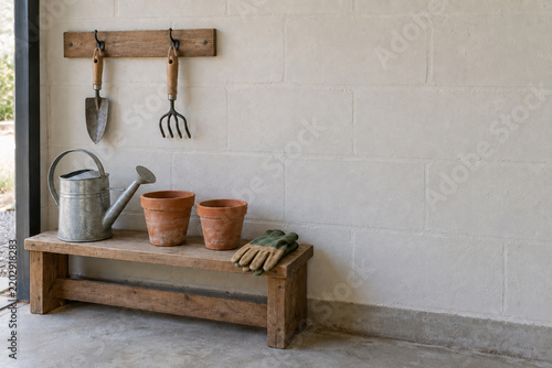 Garden tools and watering can with terracotta pots on a wooden bench against a white wall
