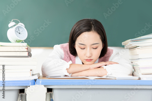 Tired Asian college student sleeping on desk with books and alarm clock in classroom