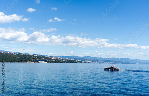 Scenic view of a tourist passenger boat cruising on the blue water of the Adriatic Sea with coastal town and mountains in the background