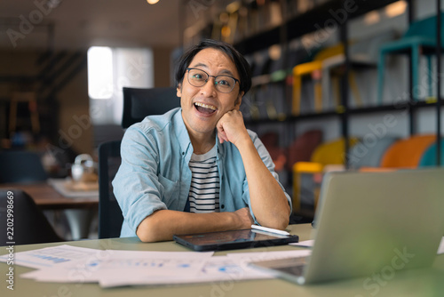 Принти на полотні Playful Asian businessman resting chin on hand at desk, daydreaming with thought