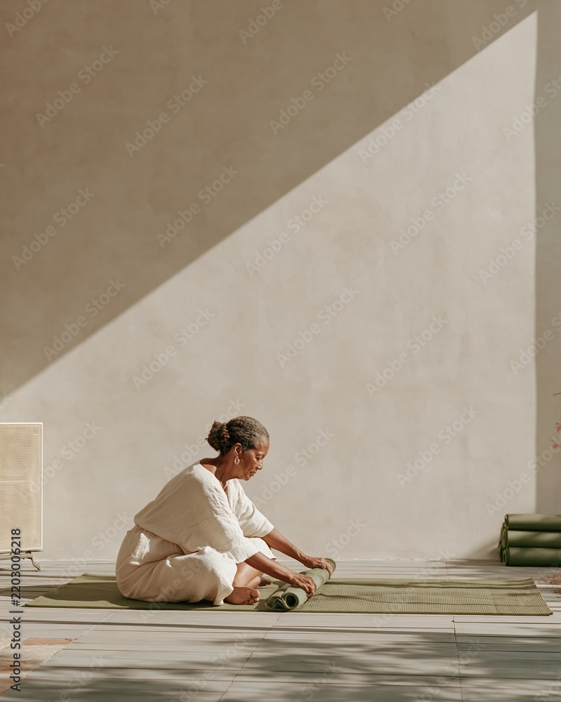 Fototapeta premium Muted cream wall with copy space and a senior Black woman unrolling a green yoga mat on a slate grey deck for a wellness festival