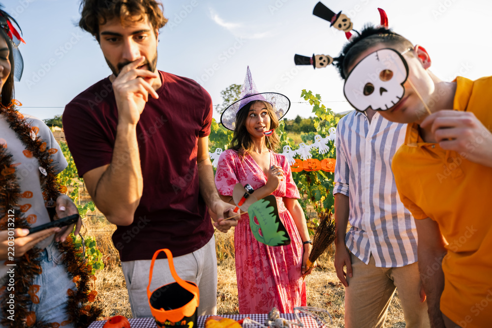 Obraz premium Young people in Halloween costumes holding skull and monster photo props at a party table.