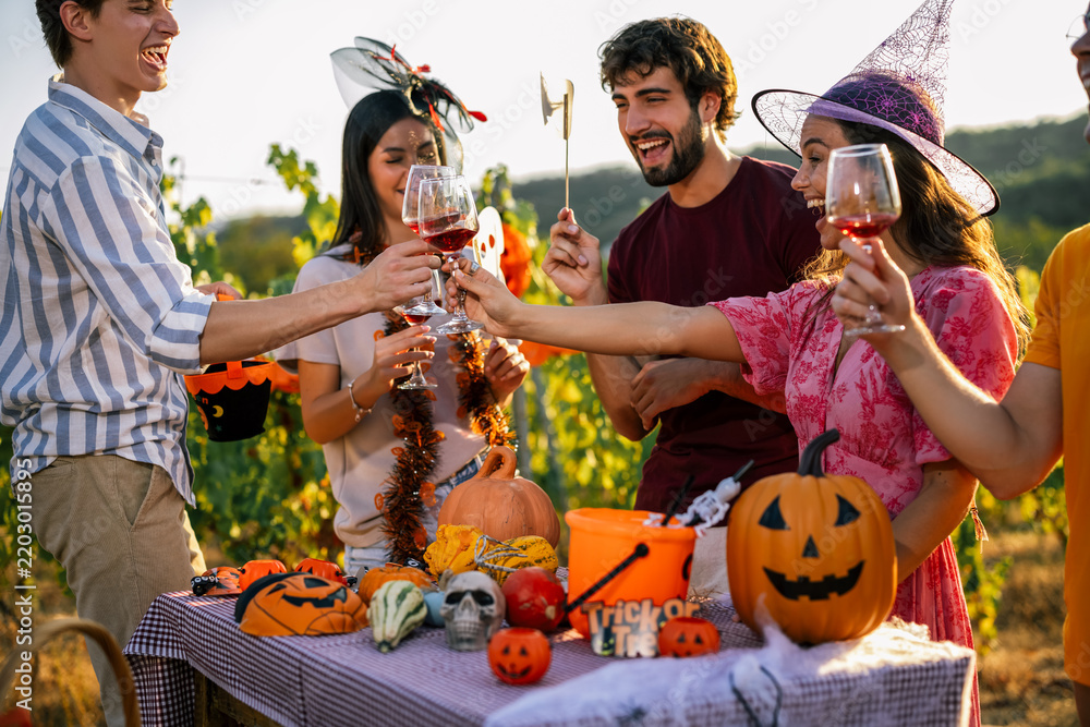 Obraz premium Group of joyful young friends wearing Halloween costumes and toasting with red wine outdoors at a vineyard party.