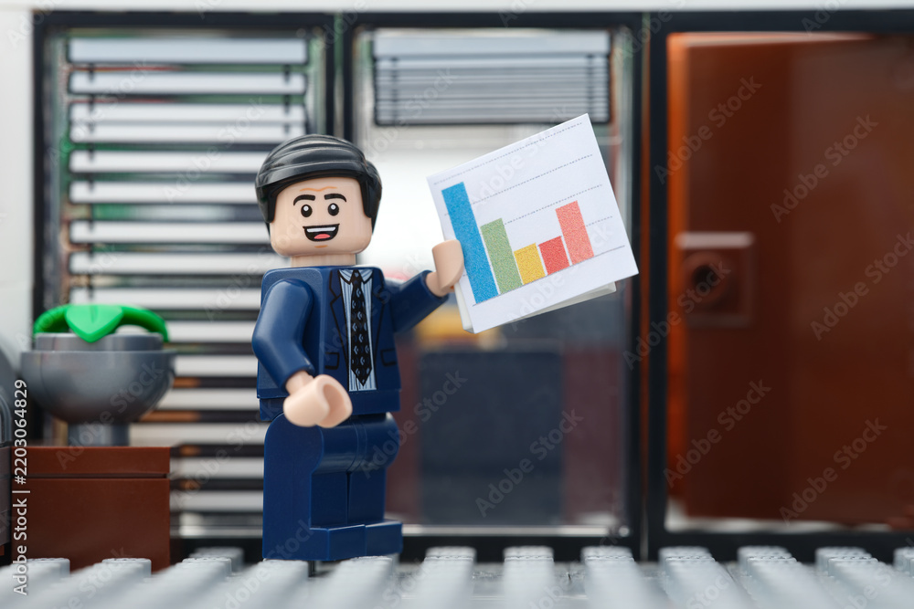 Obraz premium Tambov, Russian Federation - September 23, 2026 A happy Lego businessman minifigure standing in an office setting while holding up a colorful bar chart document.