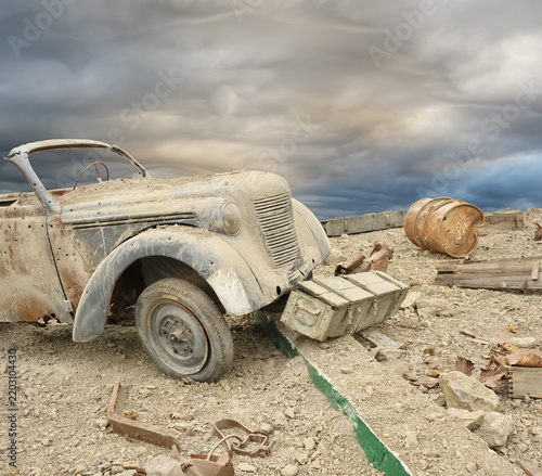 Vintage military car stands by wooden ammo box and rusty barrel on ruined battlefield under storm clouds. Dramatic historical warfare