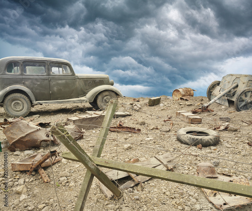 Vintage military car stands on ruined battlefield with barbed wire under dark storm clouds. Dramatic historical warfare