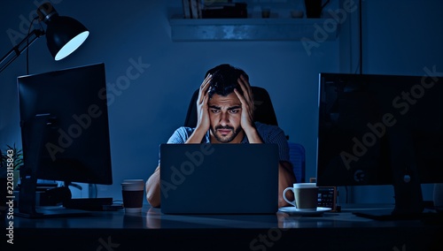 Stressed young man working late at night on a laptop in a dark office with multiple monitors