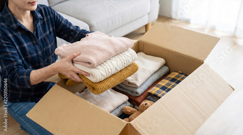 Woman packing folded sweaters and clothes into large cardboard box while organizing wardrobe at home for winter season.