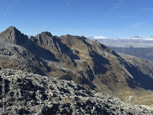 High-altitude panoramic mountain view in the Swiss Alps near Realp, Uri, showing jagged rocky peaks, dramatic ridges, alpine valleys and cloud layers under a deep blue sky in the wild landscape