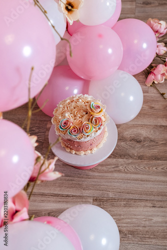 Pink birthday cake surrounded by balloons on wooden floor