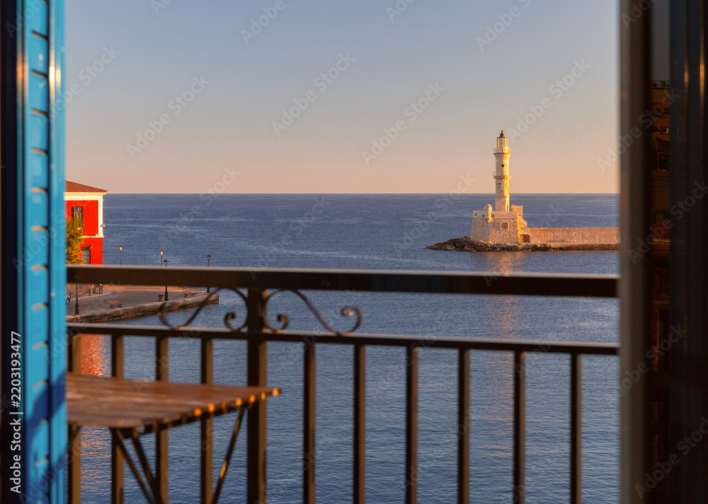 Fototapeta premium View through open balcony doors toward Chania Lighthouse, turquoise sea and boats in the Old Venetian Harbor of Chania, Crete, Greece.