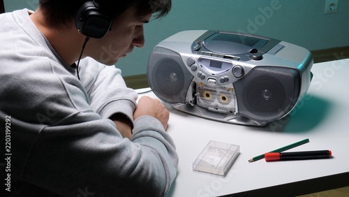 Young man wearing headphones and intently listening to music on a vintage boombox with an open cassette deck, focusing on nostalgic audio technology on a white table