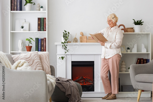 Senior woman reading book by fireplace at home