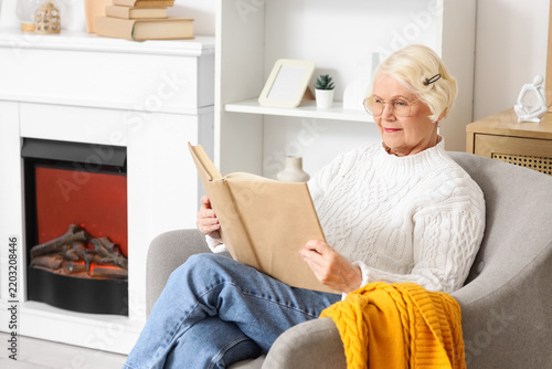 Senior woman reading book in armchair near fireplace at home
