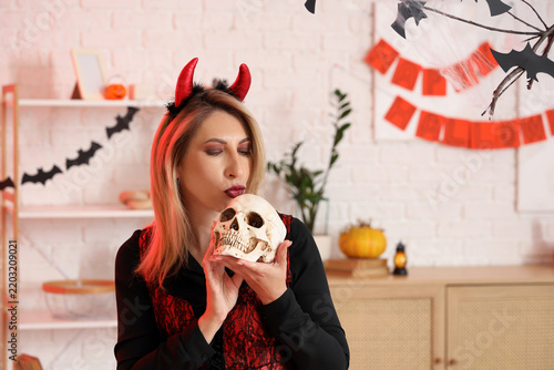 Happy mature woman dressed as devil with human skull blowing kiss at decorate...