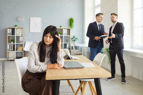 Obraz na płótnie Woman at office desk rests head in hand as peers chat by window, with workplace stress, gossip, conflict, bullying, isolation, burnout, anxiety, fatigue