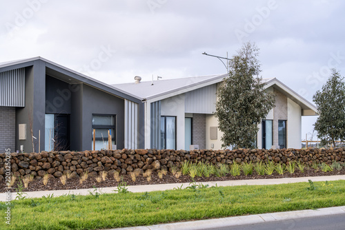Modern single-story residential townhouses behind a stacked dry-stone retaining wall, and nature strip in a new suburban housing estate in an outer suburb of Melbourne, Victoria, Australia.  