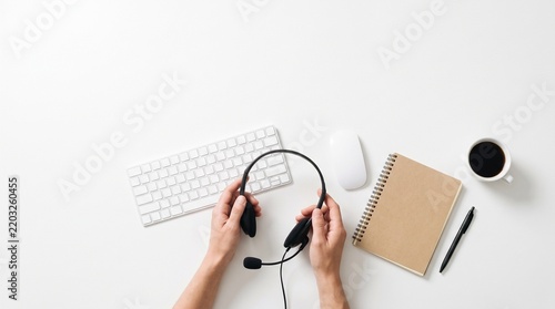 Top-down view of hands holding a headset on a white desk with keyboard, mouse, notebook, pen, and coffee mug, representing remote work or customer support.