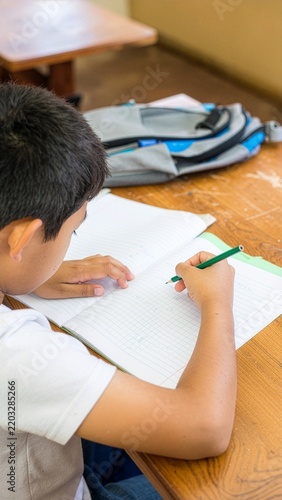 Tapety artystyczne 教室の机でノートに鉛筆で文字を書く小学生の男の子、筆箱をそばに置いて集中して勉強する学校の学習風景