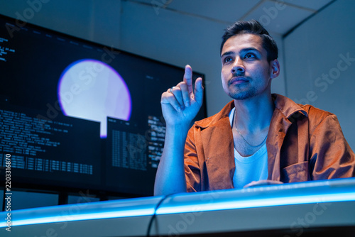 Foto A dark-haired network defence analyst concentrates at the keyboard in the dark room in a shadowy data monitoring room lit by screen glow, during an overnight monitoring shift