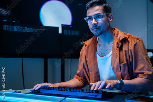 Photography A focused dark-haired cybersecurity analyst sits at the keyboard facing the camera inside a modern security operations centre after hours, securing enterprise systems