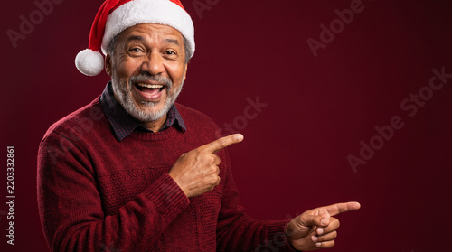 Excited mature African American man wearing Santa hat pointing to copy space against red background
