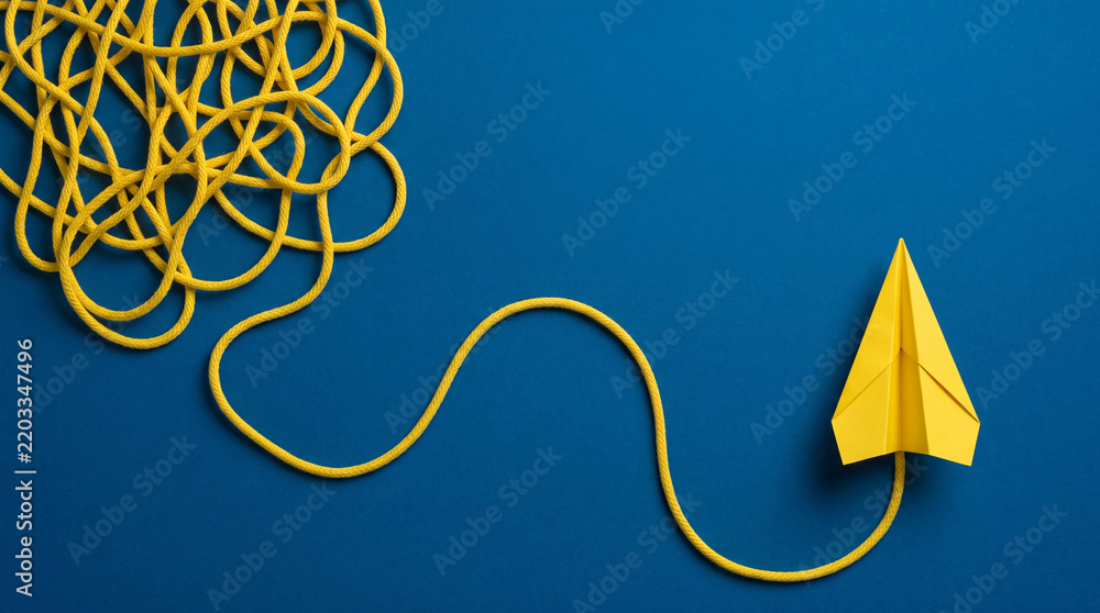 Fototapeta premium Conceptual top view of a yellow paper plane flying out from a tangled messy rope on a blue background, symbolizing finding solutions, business strategy, creative thinking, and mental clarity.