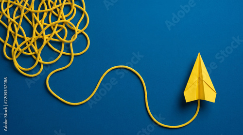 Conceptual top view of a yellow paper plane flying out from a tangled messy rope on a blue background, symbolizing finding solutions, business strategy, creative thinking, and mental clarity.