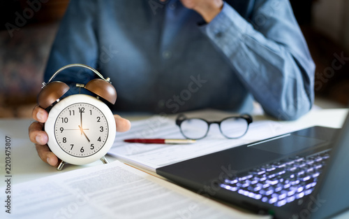 close up shot, asian businessman work in office with laptop and many documents he holding alarm clock at 5.00 PM end of work time in a day