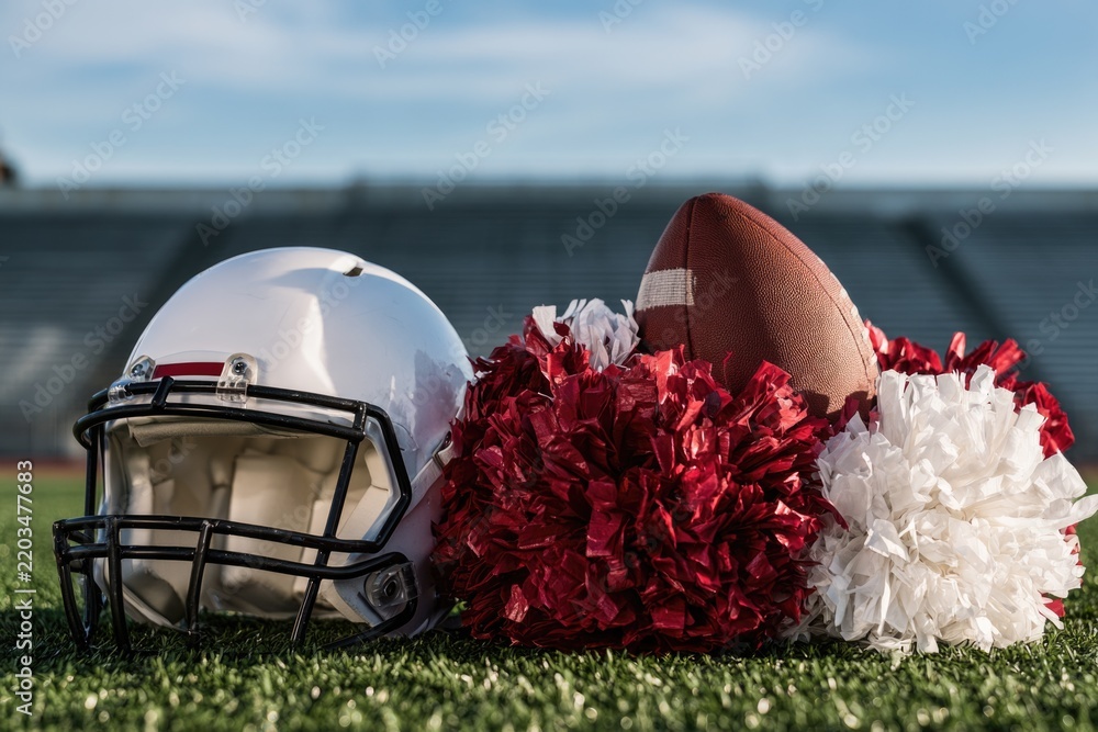 Obraz premium A white football helmet, brown football, and red and white pom-poms rest on green turf with stadium seats blurred in the background.