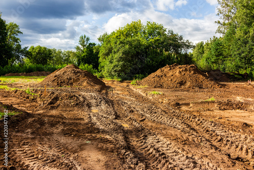 Excavation site with piles of brown soil and deep tractor tire tracks on raw ground at a summer public park during a landscape renovation project
