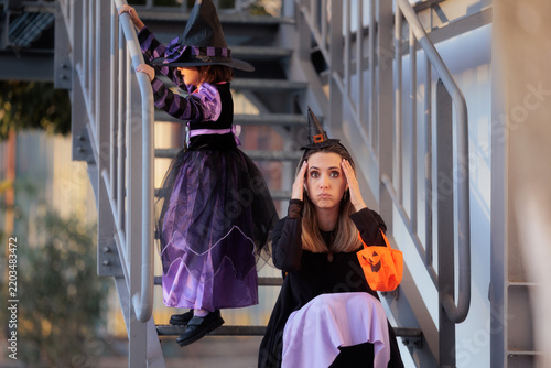 Mother Looking Tired Holding Head And Child In Witch Costume On Stairs For Halloween. Frustrated mother sitting on stairs holding pumpkin bag while her daughter climbs up in a witch costume