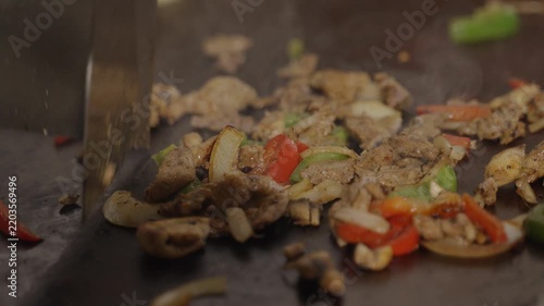 Cook chopping grilled chicken, mushrooms, onions and bell peppers on a hot flat top griddle in a busy restaurant kitchen.