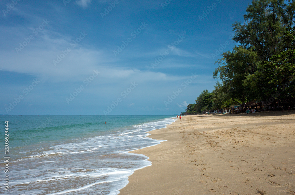 Obraz premium A peaceful scene on Koh Lanta’s Long Beach, with soft sand, gentle blue‑green waves, shaded trees and distant visitors along the shoreline