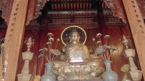 An altar with gilded bronze statue of Buddha Shakyamuni sitting in the meditation posture with vases and lotus flowers in front of the altar. Hai Duc Buddhist Institute located on Trai Thuy Hill in Nh