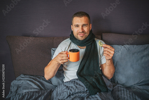 Sick but positive young man sits on bed in bedroom and looks on camer. He holds orange cup in one hand and piece of lemon in another one. Guy smiles. He looks happy.