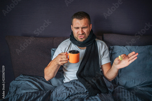 Unhappy young man sits on bed in bedroom and looks at orange cup. He holds it in one hand and piece of lemon in another one. Guy is unsatisfied. He shrinks. Liquid in cup smells disgusting.