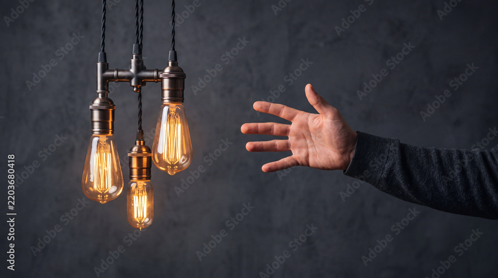 Fototapeta premium Male hand reaching towards glowing vintage Edison light bulbs against a dark textured wall background, symbolizing creativity, bright ideas, inspiration, innovation, and conceptual imagination.