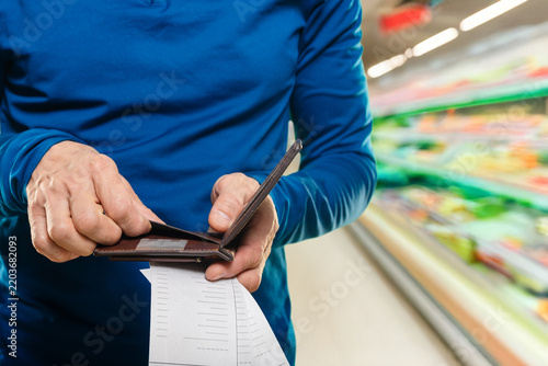 Bald man with receipts checking wallet in produce aisle, budgetconscious home cook inspecting totals while hands hold nearly empty leather wallet, fluorescent supermarket lighting,