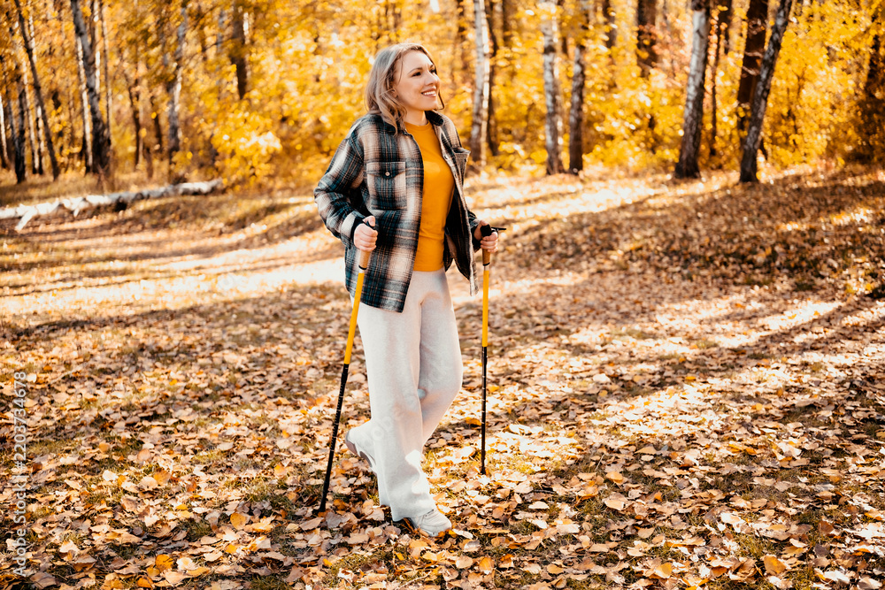 Obraz premium Active woman in autumn forest with nordic walking poles at fitness training. Fall outdoor sports and mental health improvement. Enjoying alone leisure time and escaping daily urban stress.
