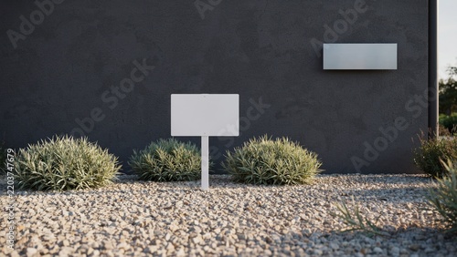 Blank white yard sign mockup erected on gravel stones beside dark building wall and green bushes
