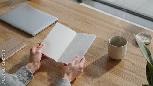 Hands opening blank bifold brochure mockup on wooden tabletop beside coffee cup in sunlight