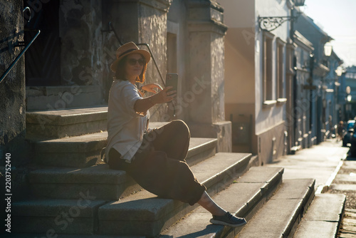 Young woman taking a selfie while sitting on historic stone steps in warm evening light. Solo traveler capturing memories with a smartphone in an old town during summer