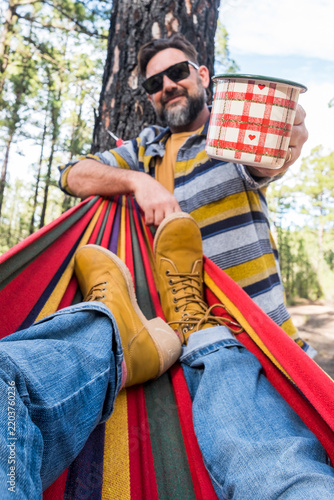 Canvastavla A bearded man offers a patterned enamel mug to a companion relaxing in a colorful hammock among pine trees, sharing coffee, laughter and outdoor camping leisure in a sunlit forest