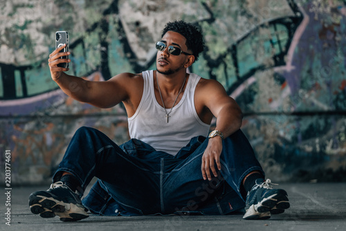 Confident young man taking a selfie with a smartphone while sitting on the ground in front of a colorful graffiti wall, cool urban street latin lifestyle portrait with a relaxed