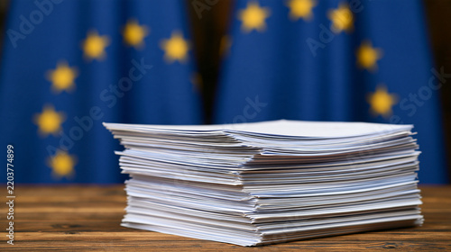 A stack of paper documents sits on a wooden desk with European flags in the background. Useful for European legislation and policy concepts.